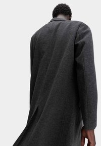 Cappotto di lana grigio scuro con design su misura, maniche lunghe, spacchetto posteriore evidente e colletto strutturato. Texture liscia e linee pulite.