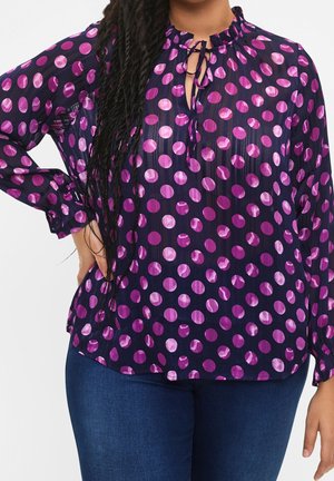 Kobieta ubrana w granatową bluzkę w różowe groszki, z długimi rękawami, falbaniaste wykończenie przy dekolcie z wiązaniem, w połączeniu z ciemnoniebieskimi jeansami.