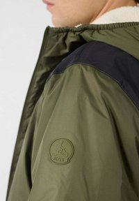 Olijfgroene jas met een zwart schouderpaneel, voorzien van een gestructureerd logopatch op de mouw en een trekkoord capuchon voor verstelbare pasvorm.