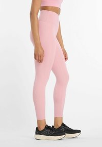Donna che indossa leggings rosa a vita alta e scarpe da ginnastica nere, in piedi di lato su uno sfondo bianco.
