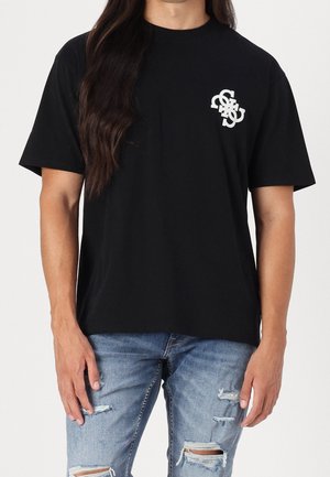 T-shirt di cotone nero a maniche corte, con un piccolo logo bianco nella zona sinistra del petto, abbinato a jeans blu strappati.