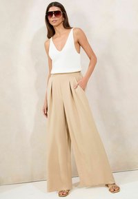Débardeur blanc à côtes associé à un pantalon beige à jambes larges et plissé. Le mannequin porte des lunettes de soleil surdimensionnées et des sandales dorées à lanières.