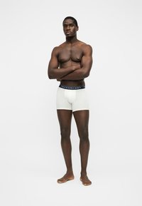 Homme musclé debout pieds nus, bras croisés, portant un boxer blanc avec une ceinture bleu marine sur un fond blanc uni.