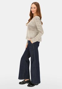 Kobieta o długich brązowych włosach, ubrana w beżowy sweter z dzianiny, ciemne szerokie dżinsy oraz czarne błyszczące mokasyny, stojąca w pozycji bocznej.