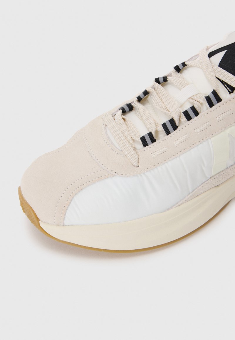Beige Sneaker mit Wildleder und glänzendem weißen Material, runder Zehenpartie, schwarzen Akzent-Schnürsenkeln und einer strukturierten Gummisohle.
