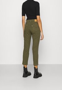 Person som bär olivgröna högmidjade jeans, svart kortärmad topp och svarta ankelboots, står vänd mot en enfärgad ljus vägg.