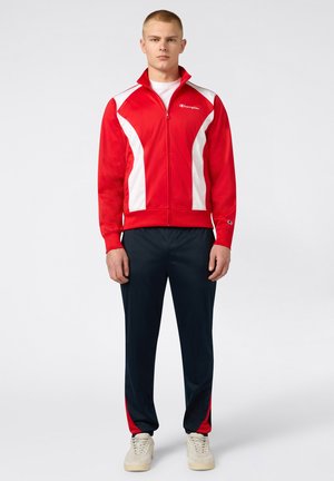 Jeune homme portant une veste de sport zippée rouge et blanche, un pantalon de survêtement foncé avec des accents rouges et des baskets blanches, debout devant un fond uni.