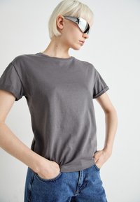 Grå bomullst-skjorte med rullede ermer, kombinert med blå denim jeans. Modellen har på seg kantede sølvsolbriller med stjernedetaljer.