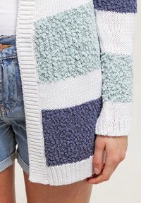 Cardigan tricoté avec des rayures texturées en bleu clair, blanc et bleu foncé. Poignets et ourlet côtelés, ouverture frontale, coupe décontractée.