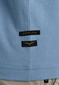 Tissu de chemise en coton bleu avec deux étiquettes noires ; l'une indique "TYPE NO : C-46" et l'autre "AMERICAN CLASSIC" avec des accents dorés.