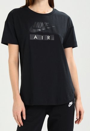Persoon draagt een zwarte Nike Air T-shirt en zwarte Nike broek met wit Nike-logo zichtbaar op de broek, tegen een effen lichte achtergrond.