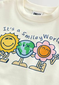 Text „It's a SmileyWorld“ nad třemi kreslenými postavičkami: usmívajícím se sluncem, Zemí a květinou držící se za ruce na krémové látce.