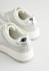 Weiße Sneakers mit silbernen Akzenten, die ein strukturiertes Mesh-Design, eine metallische Ferse und eine dicke, weiße Gummisohle aufweisen.