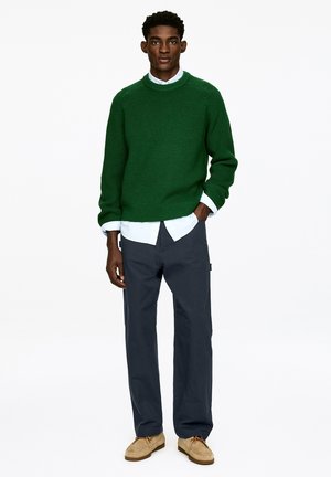 Grüner Strickpullover mit Rundhalsausschnitt, über einem hellblauen Hemd getragen. Kombiniert mit dunkler Hose und beigen Loafers. Einfaches Design, strukturierter Strick.