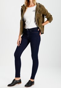 Mörkblå skinny jeans, vit t-shirt med logotyp, olivgrön jacka och svarta skor. Outfitten har en avslappnad, lager-på-lager look.