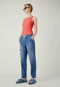 Γυναίκα που φορά black γυαλιά ηλίου, αμάνικο κοραλλί τοπ, μπλε cargo παντελόνι με κορδόνια και μπλε σανδάλια, στέκεται με τα χέρια στις τσέπες.