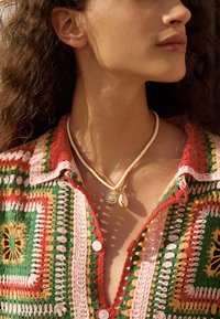 Cárdigan de crochet colorido y estampado con tonalidades rojas, verdes y rosas; collar de dos capas con un colgante de sol y un adorno de concha.