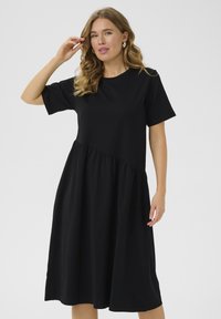 Black cotton dress with short sleeves, a round neck, and a tiered design. Soft texture with a flowy silhouette and gathered waist detail.