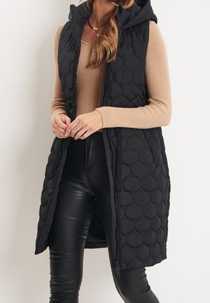 Femme portant une veste sans manches matelassée noire à capuche sur un haut beige côtelé à manches longues et un pantalon en cuir noir, posant devant un fond blanc.
