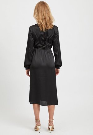 Kvinde med skulderlangt blondt hår iført en sort midi-kjole med lange ærmer og beige høje hæle, set bagfra på hvid baggrund.