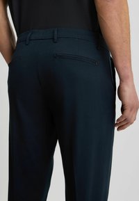 Marineblaue Hose mit glatter Textur, ausgestattet mit einem taillierten Bund, zwei Reißverschlusstaschen auf der Rückseite und einem taillierten Schnitt.