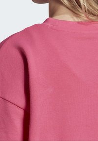 Růžová mikina s jemnou texturou, žebrovaným límcem a volným střihem. Viditelné švy a hladký povrch látky vytvářejí neformální vzhled.