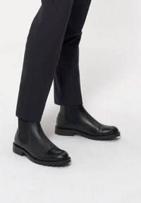 Czarne skórzane botki Chelsea z elastycznymi bocznymi panelami, okrągłym noskiem, wzmocnionym czubkiem i solidną podeszwą, noszone z dopasowanymi ciemnymi spodniami.