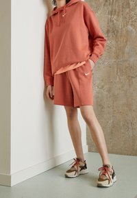 Coral hoodie with front pocket and drawstrings, paired with matching shorts. Wearing beige and brown sneakers with red laces.