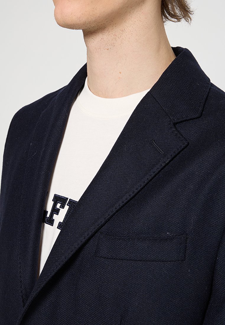 Granatowa marynarka o klapach, z teksturowanej tkaniny i pojedynczą kieszenią na piersi. Nosi się ją na jasnym t-shircie z napisem.