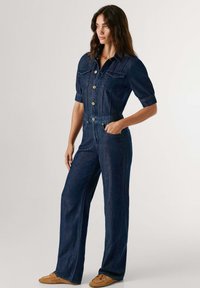 Ολόσωμη φόρμα από τζιν σε σκούρο μπλε χρώμα με εφαρμοστή μέση, κοντά μανίκια, κούμπωμα με κουμπιά, τσέπες στο στήθος και φαρδιά παντελόνα. Διακοσμημένη με χρυσά κουμπιά.