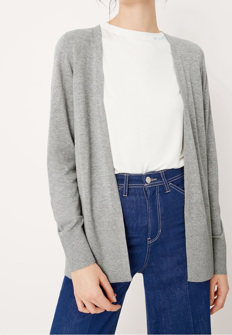 Cárdigan gris de punto con delante abierto, mangas largas, puños acanalados, llevado sobre una camisa blanca, combinado con unos vaqueros de cintura alta azules.