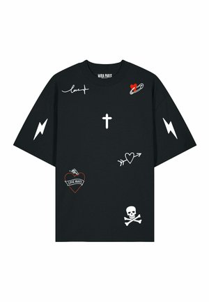 Uomo in piedi che indossa una t-shirt nera oversize con simboli grafici bianchi e rossi, pantaloni cargo grigi e sneakers bianche su sfondo semplice.