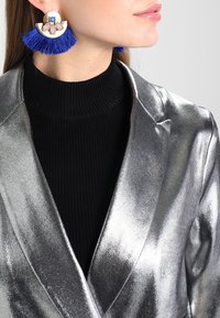 Stříbrný metalický blazer s hladkou texturou, doplněný velkými modrými třásněmi na náušnicích s zlatým a bílým květinovým detailem.