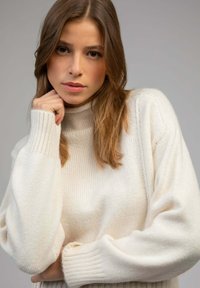 Pull en maille couleur crème avec un col montant, des poignets côtelés et une coupe décontractée, mettant en valeur une texture douce et des coutures détaillées.