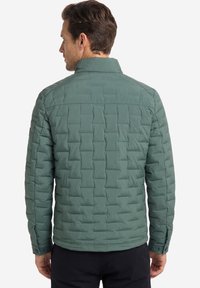 Mann trägt eine grüne gesteppte Jacke mit geknöpften Manschetten, von hinten vor einem schlichten weißen Hintergrund gezeigt.