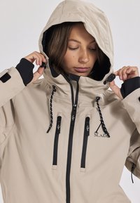 Beige wasserdichte Jacke mit Kapuze, schwarzen Akzenten, verstellbarem Zugband und mehreren Reißverschluss-Taschen, mit einem Logo auf der Brust.