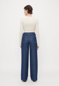 Vide ben bukser i mørk denim med elastisk talje, med dobbelte baklommer og hvit søm. Komplementert med en kremfarget ribbestrikket langermet topp.
