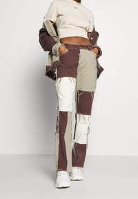 Person wearing patchwork pants and a matching jacket in brown, beige, and white with frayed edges, paired with white sneakers and a beige crop top.