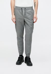 Szürke cargo nadrág elasztikus derékpánttal, karcsúsított szabással, oldalzsebekkel és diszkrét márkajelzéssel. Fekete tornacipővel párosítva.