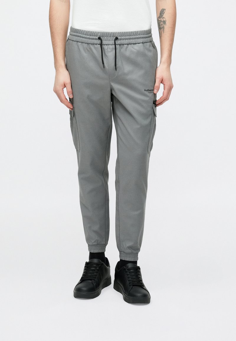 Szürke cargo nadrág elasztikus derékpánttal, karcsúsított szabással, oldalzsebekkel és diszkrét márkajelzéssel. Fekete tornacipővel párosítva.