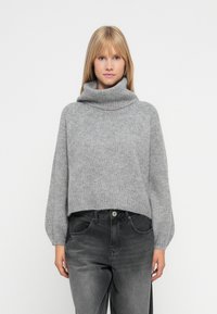 Ung kvinne iført en grå høyhalset genser og falmede svarte jeans, stående mot en ensfarget hvit bakgrunn.