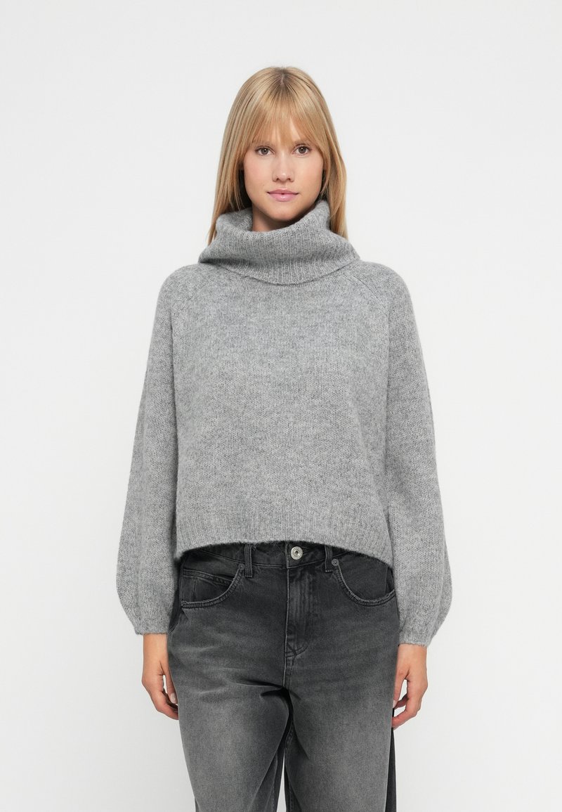 Ung kvinne iført en grå høyhalset genser og falmede svarte jeans, stående mot en ensfarget hvit bakgrunn.
