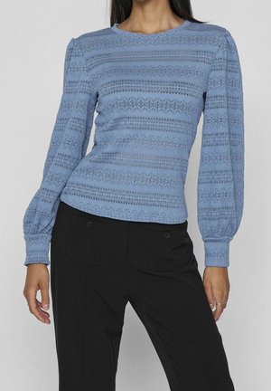 Woman wearing a light blue, long-sleeve textured knit top with black pants, standing against a plain light background.