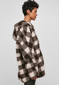 Krznena dukserica u smeđe-krem kockice s teksturiranim završetkom, opuštenog kroja i kapuljačom. Ima zatvarač sprijeda i bočne džepove.