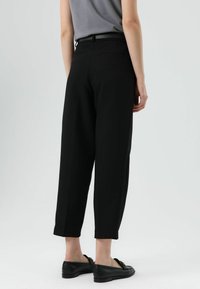Pantalon noir fuselé avec une texture lisse et des poches latérales, associé à des mocassins noirs. La ceinture présente un détail subtil de ceinture.
