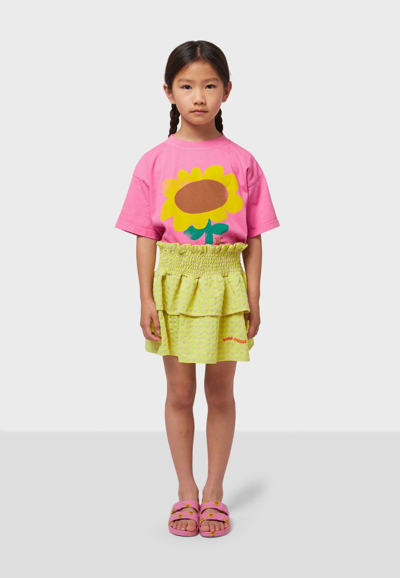 Pink T-shirt featuring a sunflower graphic. Layered yellow checkered skirt with a smocked waistband. Pink slip-on sandals with flower details.