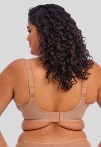 Beige draadloze beha met verstelbare bandjes, een gladde textuur en een haak-en-oogsluiting aan de achterkant. Biedt rugbedekking en ondersteuning.