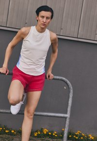 Ljusbeige ärmlös träningströja med blå detaljer och röda shorts. Vita sneakers och en grå metallräck i bakgrunden.