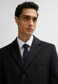 Cappotto in misto lana di colore scuro con colletto a revers, chiusura a un bottone e taschino sul petto. Indossato sopra una camicia bianca e una cravatta blu navy.