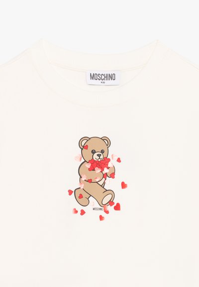 Chemise enfant couleur crème avec un ours en peluche tenant et entouré de cœurs rouges, étiquetée Moschino Kid sur le col et le motif.